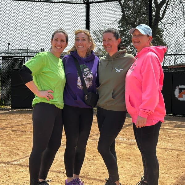 Four women in casual athletic wear smiling on a dirt field with a chain-link fence behind them.