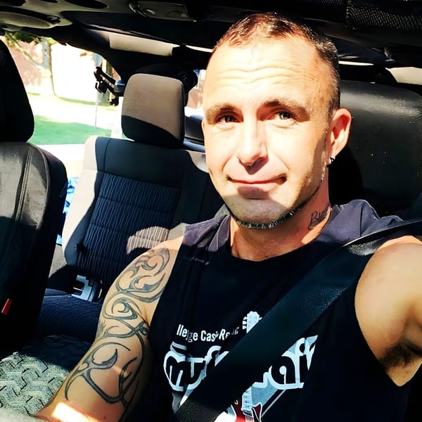 A man with tattoos on his arm and neck, wearing a black tank top, sitting in a car.