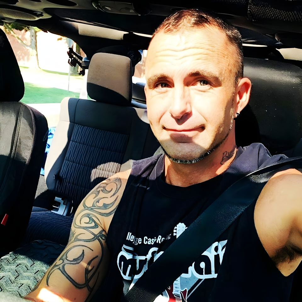 A man with tattoos on his arm and neck, wearing a black tank top, sitting in a car.