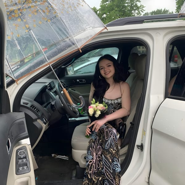 Smiling woman in a patterned dress holding a bouquet, sitting in a white car with an umbrella.