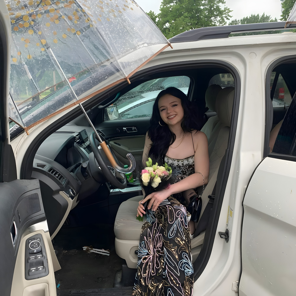 Smiling woman in a patterned dress holding a bouquet, sitting in a white car with an umbrella.