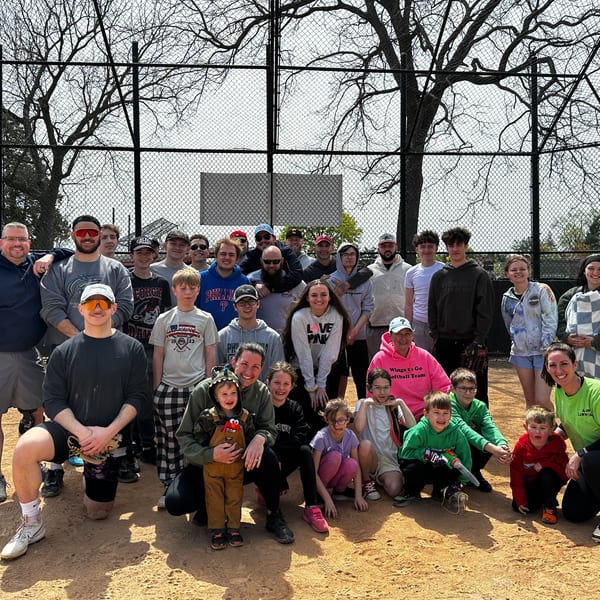 A large group of adults and children pose on a dirt baseball field in front of a chain-link fence.