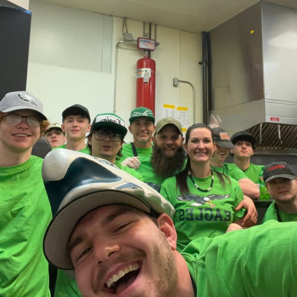 A group of smiling restaurant staff in green Eagles shirts and hats pose for a selfie in a kitchen.