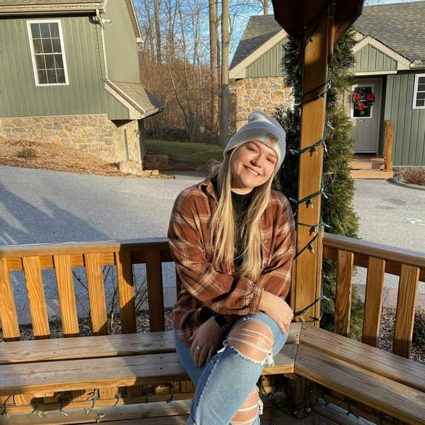 A smiling woman in a plaid shirt and beanie sits on a wooden bench outdoors.