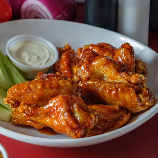 A white plate of glossy, saucy chicken wings with celery sticks and a cup of ranch dressing.