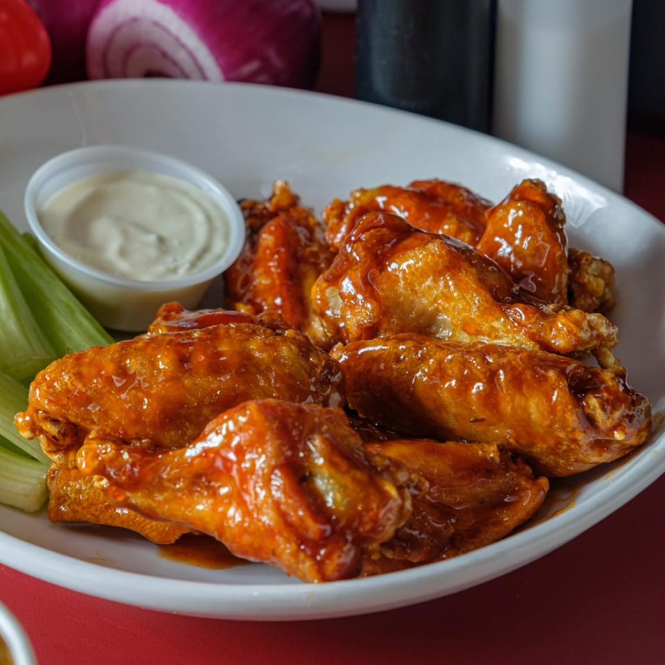 A white plate of glossy, saucy chicken wings with celery sticks and a cup of ranch dressing.