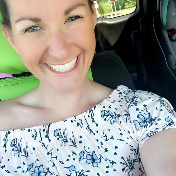 A smiling woman with blue eyes and a floral top in a car, with a child's car seat visible.
