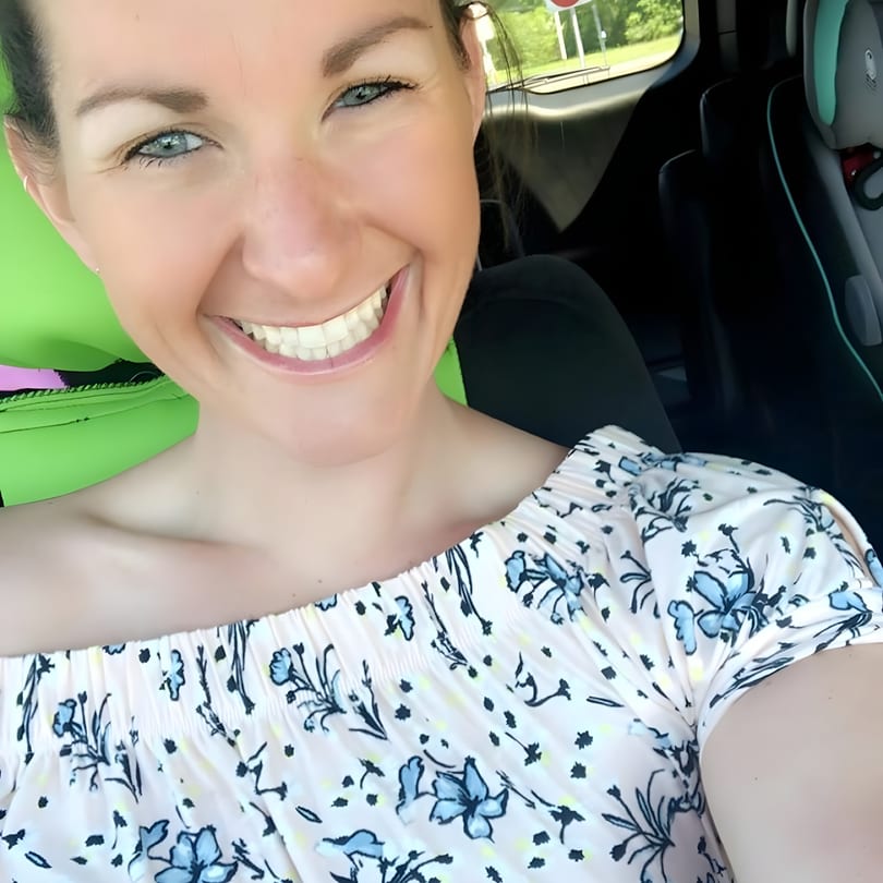 A smiling woman with blue eyes and a floral top in a car, with a child's car seat visible.