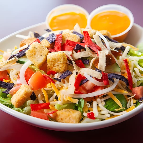 A vibrant salad with lettuce, tomatoes, onions, cheese, croutons, and colorful tortilla strips, with two cups of dressing.
