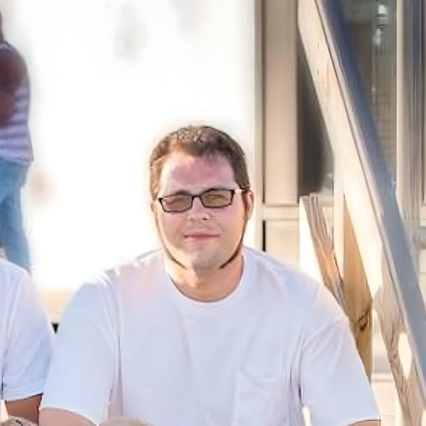 A man in glasses and a white t-shirt sits outdoors, looking directly at the camera.