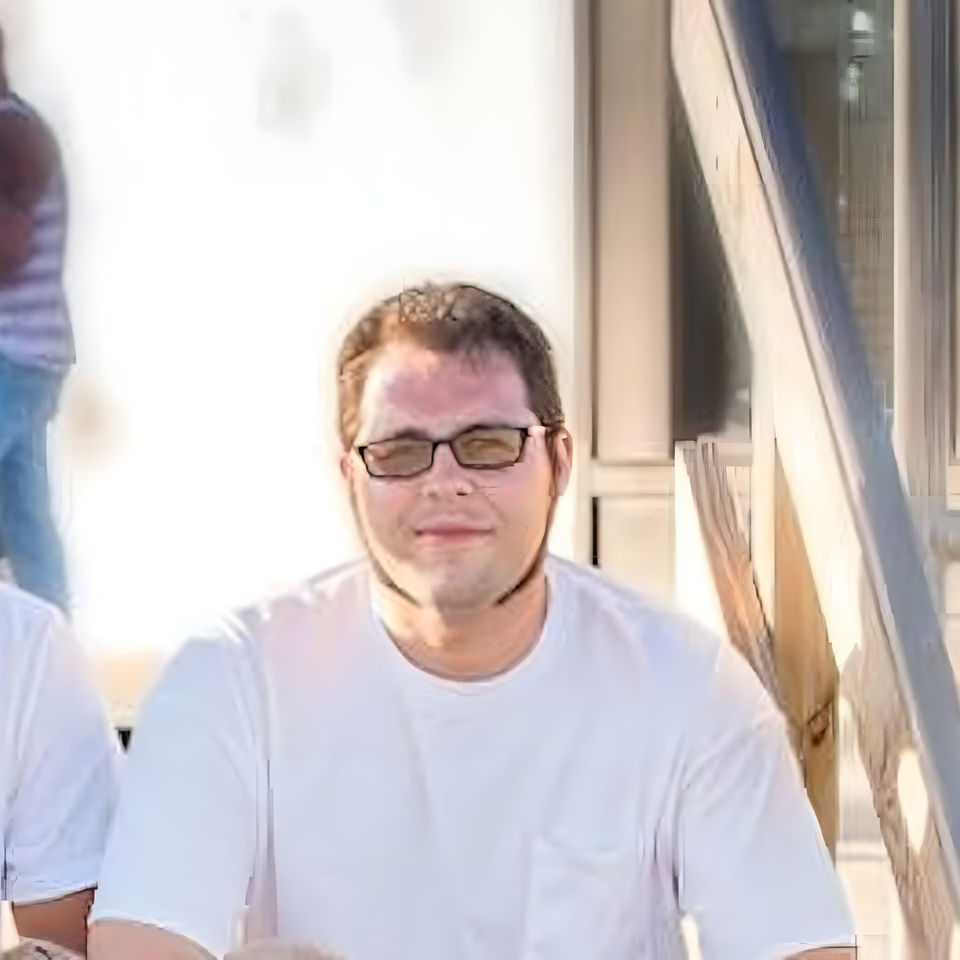 A man in glasses and a white t-shirt sits outdoors, looking directly at the camera.